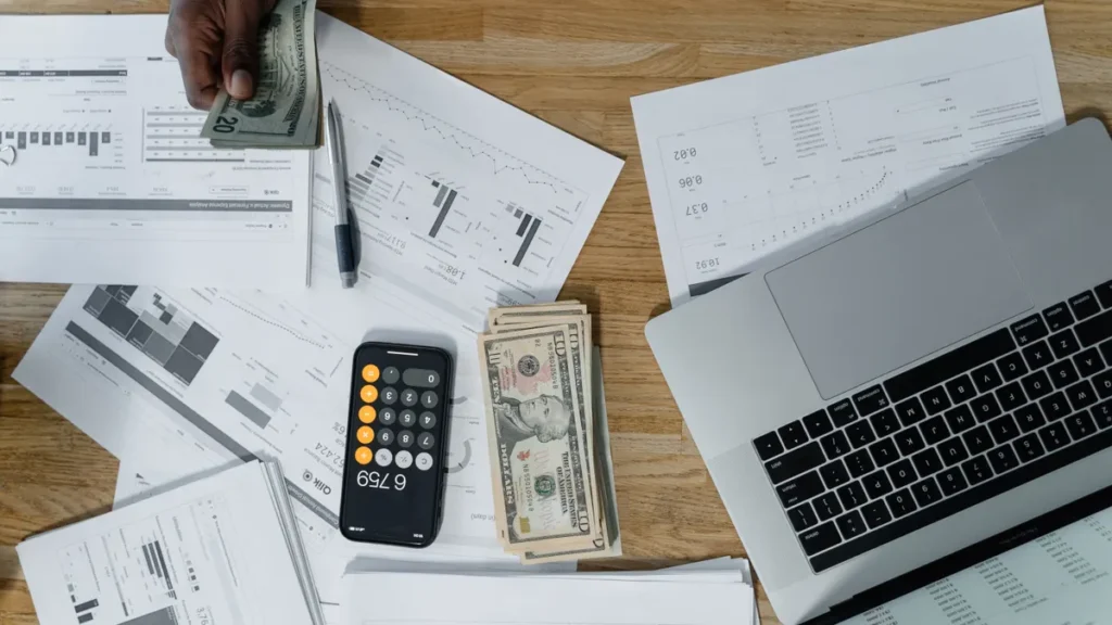 Desk covered with pay records, cash, a phone calculator and a laptop for tracking year-to-date earnings