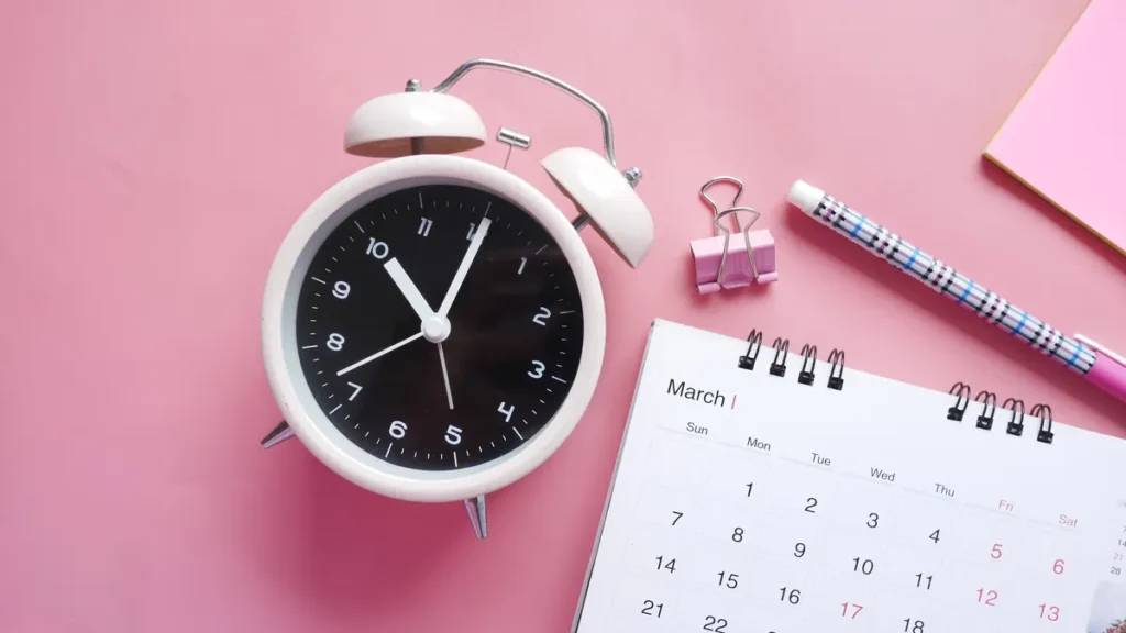 Alarm clock beside a monthly calendar on a pink desk, a reminder to track hours and time off